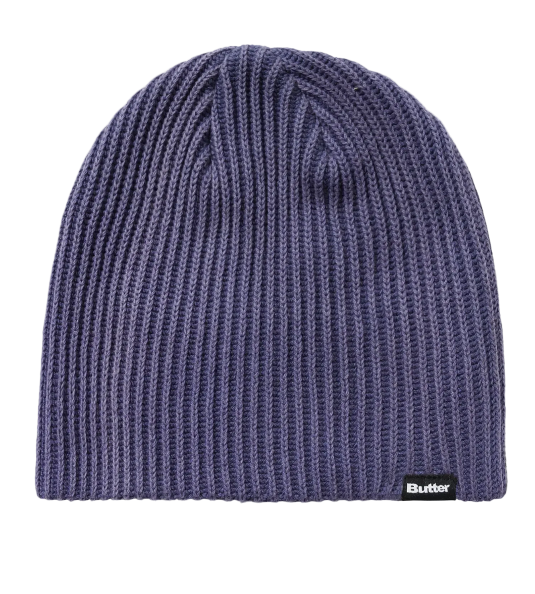 A ribbed knit beanie hat in a muted periwinkle or dusty purple color, featuring a small black brand patch on the lower right edge with the text 'Butter'. 